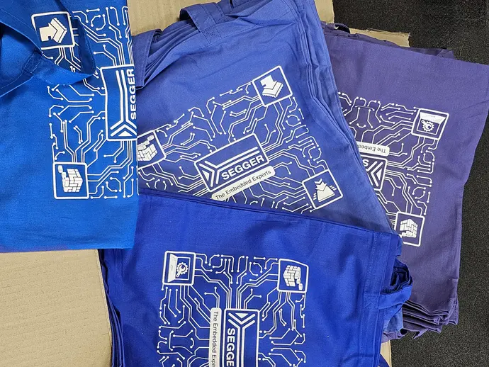 A stack of colored bags in blue and purple features a white graphic design of circuit patterns. The bags display the SEGGER logo and the phrase "The Embedded Experts," along with various icons representing technology and embedded systems.