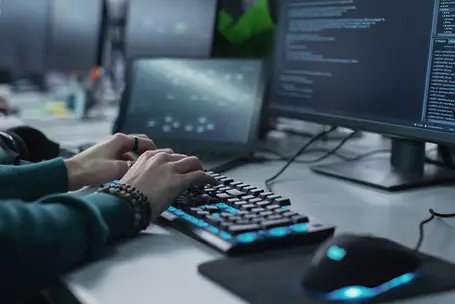 A close-up view of hands typing on a mechanical keyboard, with a computer mouse nearby. The background features multiple computer screens displaying lines of code, indicating a programming or software development setting.