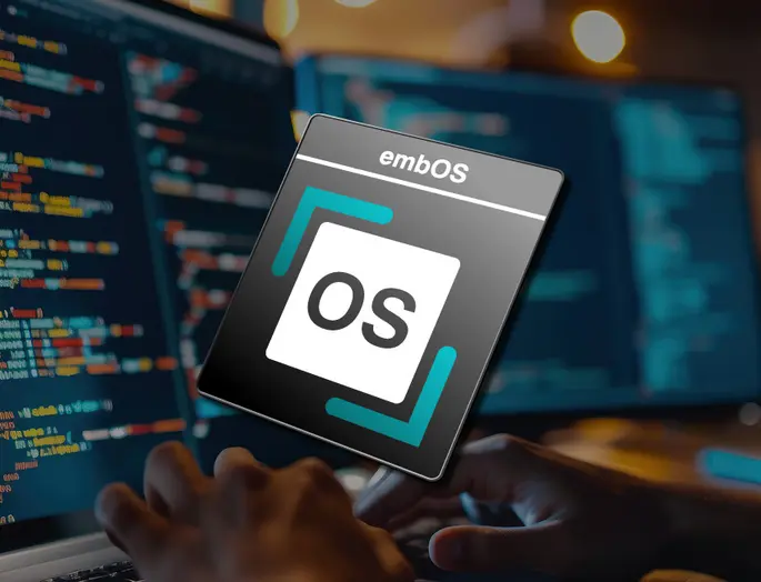 A close-up of hands typing on a keyboard, with a prominently displayed logo of "embOS," featuring the letters "OS" in a square, surrounded by a colored border. In the background, blurred code appears on screens, indicating a programming or tech environment.