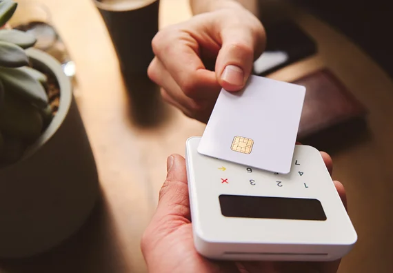 A hand is holding a credit card above a small card reader on a table. The card reader has buttons and a display screen. The background features a succulent plant in a pot and a coffee cup.