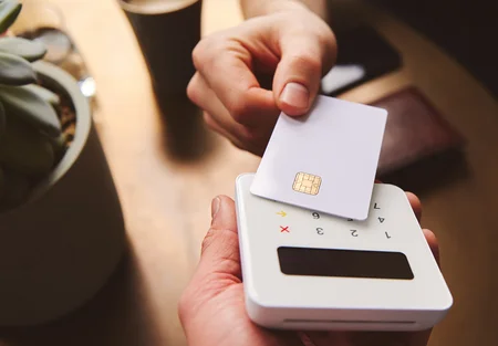 A hand is holding a credit card above a small card reader on a table. The card reader has buttons and a display screen. The background features a succulent plant in a pot and a coffee cup.