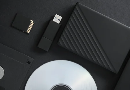 A collection of data storage devices arranged on a dark background. It includes a black external hard drive, a USB flash drive, a microSD card, a CD, and a floppy disk, showcasing various formats for digital storage and transfer.