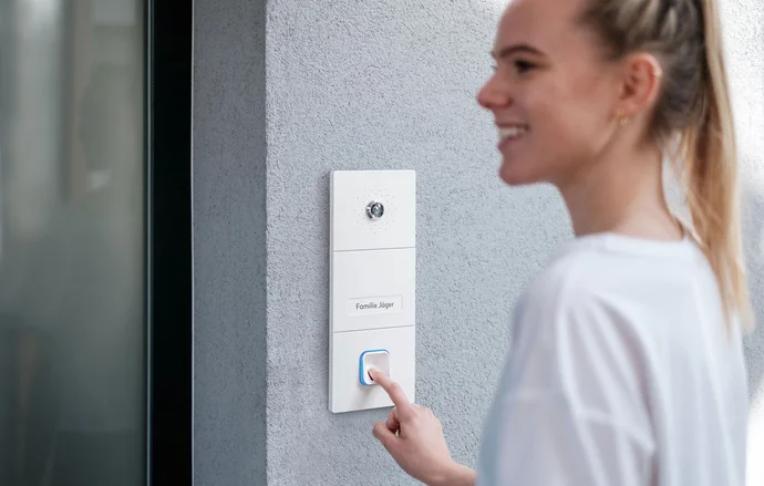A young woman is smiling and pressing the button on a modern intercom system labeled "Familie Jäger," mounted on a gray wall. The intercom features a small camera above the button, indicating it's used for communicating with visitors.