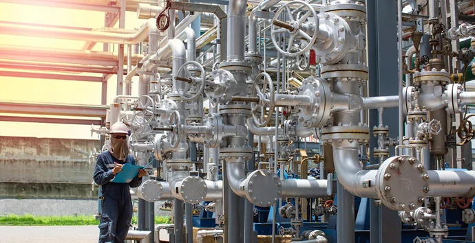 A worker in a safety helmet and protective clothing inspects complex pipelines and valves in an industrial facility. He is holding a clipboard and appears to be taking notes or monitoring equipment in a well-lit environment.