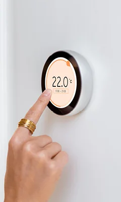 A close-up of a hand adjusting a modern thermostat displaying a temperature of 22.0°C. The thermostat features a circular design with a digital screen showing the current setting and schedule.
