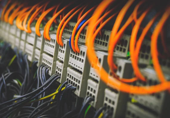 A close-up image of a row of network switches with multiple colorful cables connected. Prominent orange cables stand out among other black and yellow wires, illustrating a high-tech networking setup. The background is slightly blurred, emphasizing the equipment in focus.