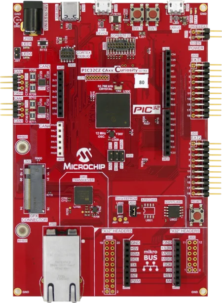 A red circuit board labeled "Microchip PIC32CZ CAXX Curiosity Ultra," featuring various electronic components, ports, and connectors including USB, power supply, and mikroBUS headers. The board includes a PIC32 microcontroller and is designed for development and prototyping purposes.