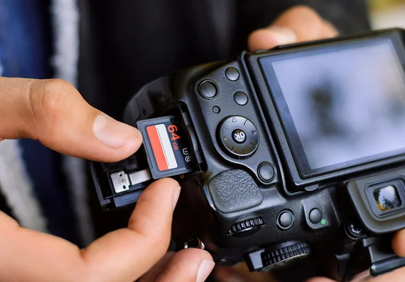 A person's hand inserts a 64GB memory card into the side slot of a digital camera. The camera has various buttons and a screen visible, indicating it is being prepared for use.