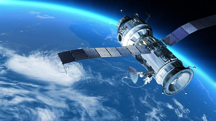 A spacecraft or satellite orbits above Earth, showcasing solar panels and communication antennas. The background features blue skies fading to darker space, with visible clouds on the planet's surface below, highlighting the satellite's position in the vastness of outer space.
