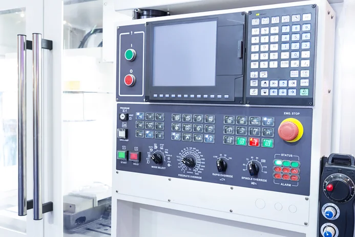 A close-up view of a machine control panel featuring various buttons, knobs, and a digital display. The controls include emergency stop, program selection, spindle override, and feed rate settings, designed for operating a CNC machine or similar equipment.