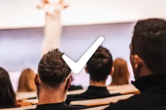 A blurred image shows the backs of several people in an audience, focused on a speaker in the foreground, who is partially visible. A large checkmark graphic overlays the scene, suggesting approval or a positive message in the context of a presentation.