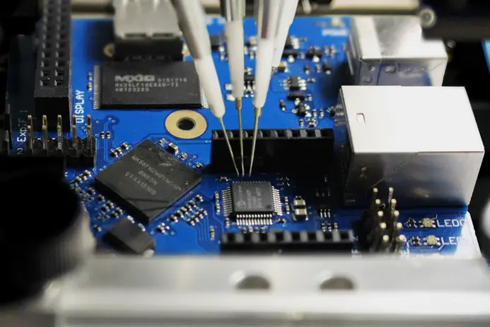 Close-up view of a blue printed circuit board featuring multiple electronic components, including chips and connectors. Three probing tools are seen making contact with the circuit, indicating a testing or diagnostic process. The setting suggests a technical or engineering environment.