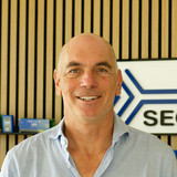 A smiling man with short hair stands in front of a wooden wall featuring a SEGGER logo. Behind him are several boxes of electronic devices displayed on shelves. He is wearing a light blue shirt and appears to be in a professional setting.