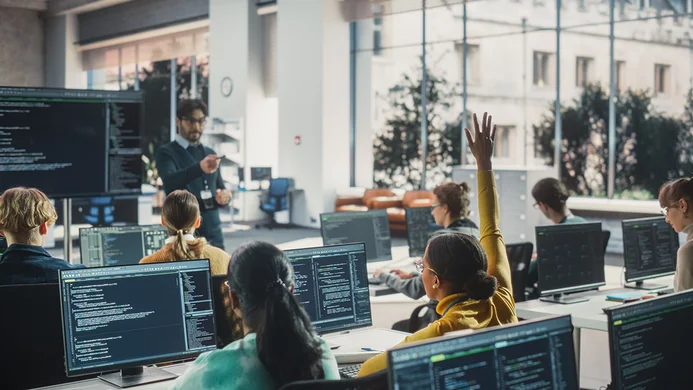 A classroom setting featuring a teacher at the front, engaging students seated at desks with computers. Some students appear to be actively participating, with one raising a hand. Multiple screens display coding and programming content, highlighting a tech-focused learning environment. Large windows allow natural light in.