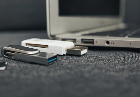 A black USB drive and a white USB flash drive are positioned near the edge of a laptop. The laptop's ports are visible in the background, indicating a workspace setting focused on data storage and transfer.