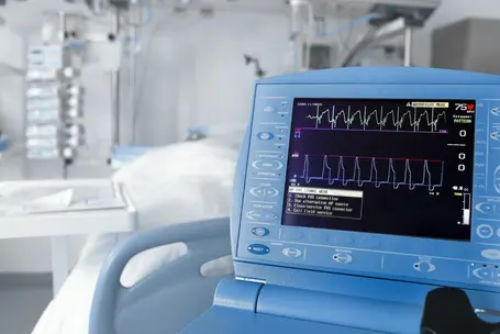 A close-up of a medical monitor displaying heart rate and waveform data. The screen shows green and blue graphs, indicating vital signs, while instructions for checking connections are visible. The background resembles a hospital setting with medical equipment.