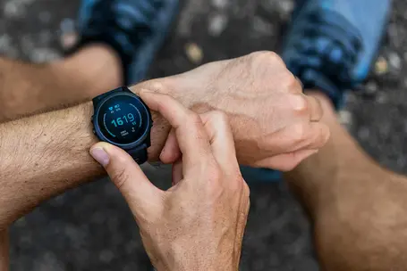 A person is checking a smartwatch on their wrist, displaying the time and various fitness metrics. The hand is positioned to interact with the watch screen, which shows numerical values related to a workout or activity.
