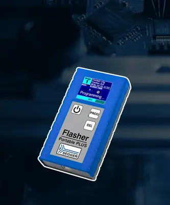 A blue and gray portable programming device labeled "Flasher Portable PLUS" is shown. It features a small screen displaying "Programming..." and a progress percentage. The device has buttons for power, programming (PROG), and selection (SEL). It appears to be used in electronics development or testing.