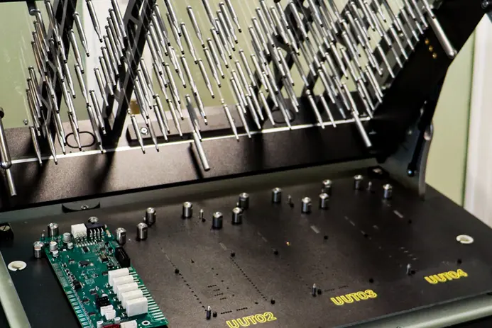 The image shows a close-up view of a testing machine with multiple metallic probes arranged in a vertical pattern. Below, a green circuit board with various components is visible. The machine has labels "UUT02," "UUT03," and "UUT04" on its surface, indicating different testing areas.