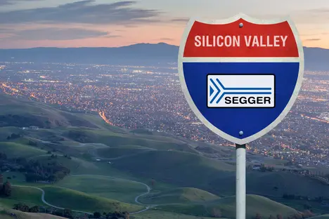 A highway sign indicating "Silicon Valley" with a logo for "SEGGER" in the center. The background features rolling green hills and a city illuminated by evening lights. The overall scene captures the transition from rural to urban landscape.