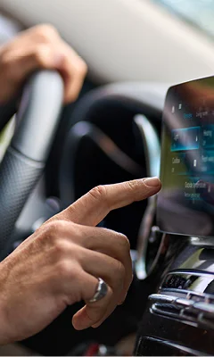 A close-up of a driver's hand interacting with a touchscreen display in a car. The hand is pointing at the screen, which shows various options and settings. The steering wheel is partially visible in the background. The focus is on the hand and the touchscreen interface.