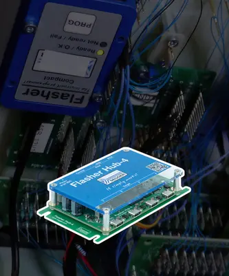 The image features a blue electronic circuit board labeled "Flasher Hub 4," prominently displayed with several connection ports on its surface. Behind it, additional circuit boards and tangled wires are visible, indicating a complex electronic setup.