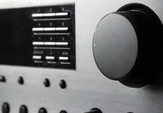 Close-up image of a silver audio control panel featuring various buttons and a prominent volume knob. The knob is round and black, with a small indicator light. The background includes additional smaller knobs for bass and treble adjustments.