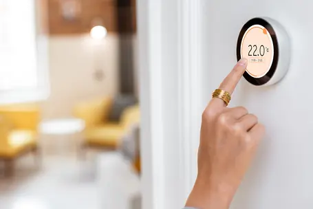 A hand is adjusting a modern thermostat displaying a temperature of 22°C. The background shows a softly lit room with yellow chairs and a small table, suggesting a cozy living space.