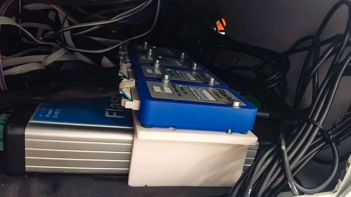 A collection of electronic equipment partially concealed by various cables. A blue control panel with five buttons and green indicator lights is positioned above a silver power supply labeled "Flash." The setup appears to be part of a larger electronic system.