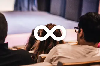 In a theater, the back of two seated individuals is visible. A symbol resembling an infinity sign is overlaid in the center, suggesting themes of continuity or limitless possibilities, amidst a blurred stage backdrop. The image conveys a sense of engagement in a performance or discussion.