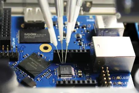 An electronic circuit board is depicted with several components, including chips and connectors. Three metal probes are positioned above the board, seemingly making contact with specific points for testing or assembly. The background is blurred, emphasizing the intricate details of the board.
