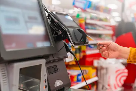 A close-up view of a card reader at a checkout counter, with a hand holding a payment card near the device. The background shows a retail environment with shelves stocked with various products.